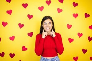 Woman wearing a red sweater smiling with a finger on each cheek, standing in front of a yellow background with red hearts on it.